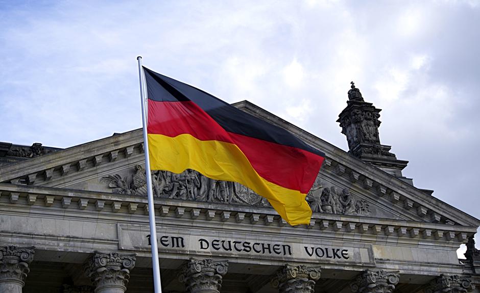 Berlin'deki tarihi Reichstag binasının önünde dalgalanan Alman bayrağı, arka planda bulutlu gökyüzünü yansıtıyor.